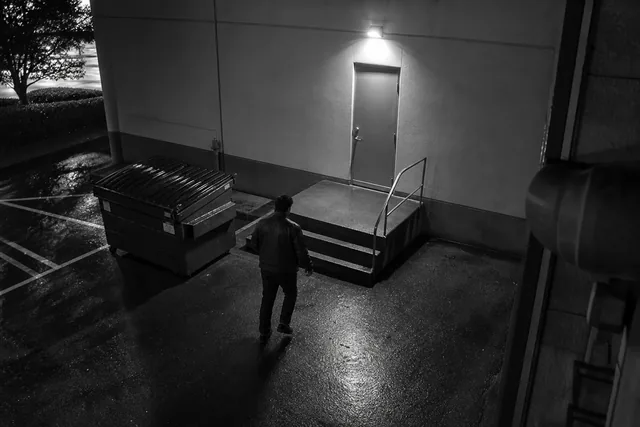 Security camera view of a building back entrance at night with a person walking toward the door
