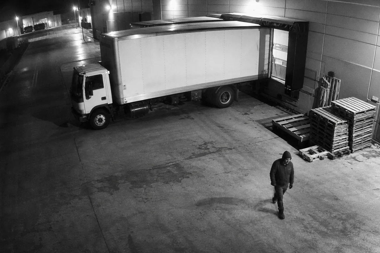 Night view from a security camera over a warehouse loading dock with a person and a parked truck