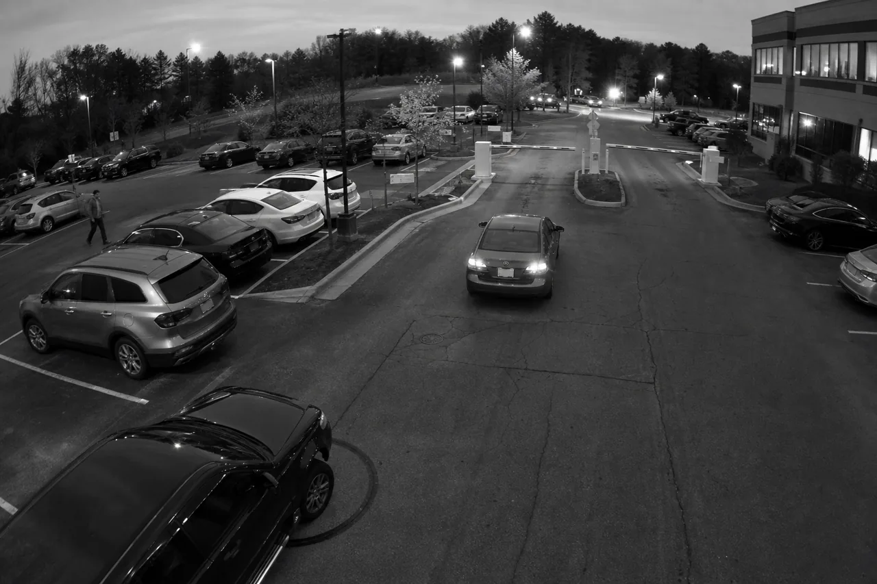 Overhead security camera view of an office parking lot at dusk with cars and a pedestrian